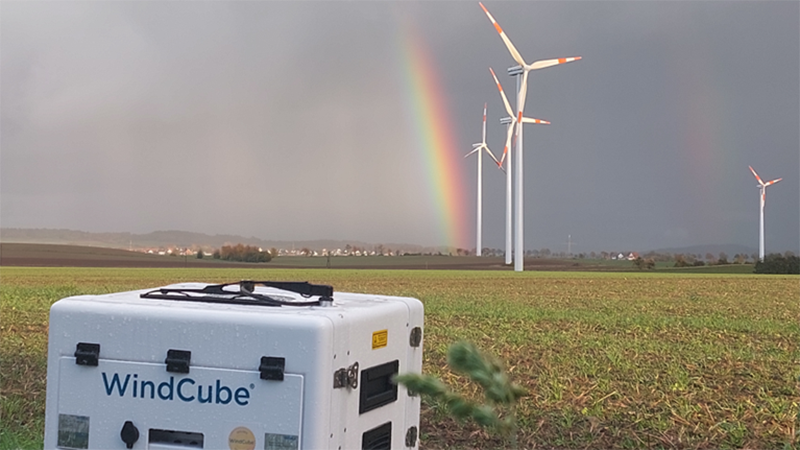 Windcube in field, wind turbines and rainbow in background